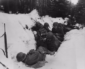 U.S. infantrymen of the 9th Infantry Regiment, 2nd Infantry Division, First U.S. Army, crouch in a snow-filled ditch, taking shelter from a German artillery barrage during the Battle of Heartbreak Crossroads in the Krinkelter woods on 14 December 1944.