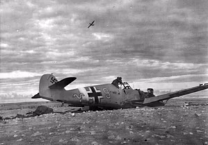 A damaged German Messerschmitt Me 109F, yellow 9 of III/JG27 at Daba, Egypt, in November 1942. An RAF Hawker Hurricane flying overhead.