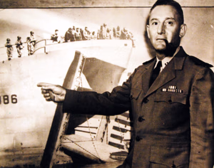 80-G-46566: Lieutenant Harold R. Fleck, USNR, poses against a photograph of his ship USS LST 386, as he recalls how the giant landing craft underweight the experiments which made it the first “junior aircraft carrier.” The event took place in North Africa. A light flight runway had been installed aboard the LST, “the plane took off like a bird leaving a tree,” Lieutenant Fleck says. He described the first combat use of the LST as a “carrier” during the Sicilian landings at Licata as a “matter of routine.” Released September 19, 1944. U.S. Navy Photograph, now in the collections of the National Archives. (2016/06/07).