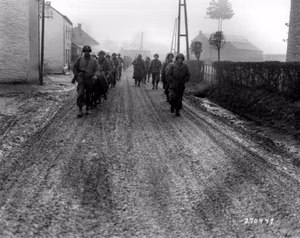U.S. troops of the 28th Infantry Division, who have been regrouped in security platoons for the defense of Bastogne, Belgium, march down a street in Bastogne. Some of these soldiers lost their weapons during the German advance in this area. Signal Corps Photo #ETO-HQ-44-30380