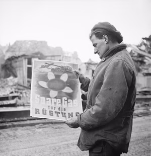 A soldier standing in the ruins of Kleve , reading a German poster calling for civilians to conserve fuel supplies for the war effort.