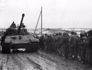 A German Tiger II tank from Schwere SS-Panzer Abteilung 501 passes a column of GIs from the 99th Infantry Division captured near Honsfeld and Lanzerath, Belgium, during the opening day of the Battle of the Bulge, 16 December 1944. The town of Merlscheid, Germany, is in the background.