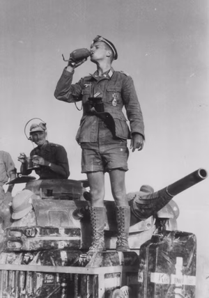 Cropped version of A German Tank Commander Stands Atop His Tank Drinking From a Canteen National Archives Catalog, National Archives and Records Administration , U.S.A.): 242-GAP-190B-4 001 WW2 (Record Group 242: National Archives Collection of Foreign Records Seized / Series: Photographs taken by Propaganda Units of the German Armed Forces (Wehrmacht) and the Waffen-SS) A World War II German officer in a Wehrmacht Heer 'Panzer' division from the Afrika Korps drinks from a canteen while standing atop a PzKpfw III tank in early 1941. He wears a tropical uniform with shorts and lace-up boots. He is awarded the Iron Cross and the Panzerkampfabzeichen . A pith helmet and jerry cans for carrying water are visible on the tank. In the turret of the tank, a comrade is seen with a headset, eating from his mess kit.