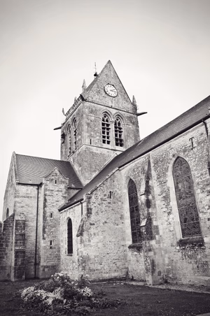 The Sainte-Mère-Église church in Normandy was built in the 11th century in the Romanesque style and completed four centuries later in the Gothic style. It’s topped by a saddleback bell tower and gained a place in history on D-Day as one of the first towns to be liberated. This was achieved through the efforts of American paratroopers who were dropped into Sainte-Mère-Église in the early hours of 6th June 1944. Photo from the site of Airborne Museum , a French museum dedicated to the memory of paratroopers of the 82nd and 101st Airborne Divisions of the United States Army who parachuted into Normandy on the night of June 5–6, 1944. Sainte-Mère-Église, Arrondissement de Cherbourg, Normandy. Summer holidays in France, August 2019