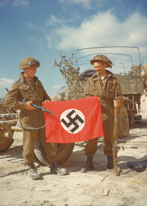 Ces deux soldats canadiens arborent un drapeau nazi qu'ils ont capturé dans les galeries de la carrière des Aucrais à Haut-Mesnil. Le fanion nazi ne semble pas avoir beaucoup servi et était vraisemblablement plié dans la sacoche de l'attelage, celle qui gît à leurs pieds sur la droite de la photo. Derrière les deux soldats, ce qui ressemble à une remorque "polyvalente" allemande Sd Anh 104, voir ici : www.lonesentry.com/ordnance/sd-anh-203-special-trailer.html Voir le camouflage fait de branches accrochées sur les côtés du camion. Le soldat de gauche est muni d'une Sten MkII fr.wikipedia.org/wiki/Sten_Mk_II et celui de droite d'un fusil Lee-Enfield n°4 MkI fr.wikipedia.org/wiki/Lee_Enfield_n%C2%B04 Photo prise le 10 Août 1944. A gauche le Pte. L.B. Roy, Cornwall, Ontario du The Queen's Own Cameron Highlanders of Canada de la 6th Infantry Brigade de la 2nd Cdn ID, il porte sur son casque une paire de lunettes de motocycliste allemand, voir ici: www.flickr.com/photos/mlq/8500700876/in/photostream Voir ici, avec un autre camarade au même endroit : www.flickr.com/photos/mlq/2547930365/in/pool-autresphotos... www.flickr.com/photos/mlq/2547930361/ Ce hameau a été libéré le 8 août, dans le cadre de " Operation Totalize " par The Argyll and Sutherland Highlanders of Canada-Princess Louise's de la 10th Inf. Brigade de la 4th Canadian Armoured Div. Dans les galeries de la carrière des Aucrais, les Allemands ont stocké du matériel, et une maquette de V2 à l'échelle 1 avec un chariot de transport du missile. Le 14 août, une erreur d'interprétation de signaux de fumée, fait que les bombardiers lourds du Bomber Command larguent leurs bombes sur les Canadiens et les Polonais à Haut-Mesnil faisant 400 victimes dont 65 morts. Pour aller plus loin : fr.wikipedia.org/wiki/Op%C3%A9ration_Totalize www.etab.ac-caen.fr/discip/geologie/mesozoi/bathoni/calca...