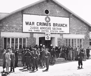 Spectators and U.S. Army military police wait outside of the building where 74 SS men will be sentenced for their roles in perpetrating the Malmedy atrocity.