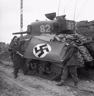 A Sherman tank crew from 27th Armoured Brigade with a German swastika flag they captured during the attack on Caen, Normandy, 10 July 1944. Sherman tank crewmen L/Cpl S James and Sgt H Coe of 27th Armoured Brigade hold up a German swastika flag they captured during the attack on Caen, 10 July 1944.