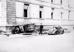 RAAF photographer Sergeant Douglas George Cracknell standing outside the damaged Reichskanzlei building in Berlin, Germany. Note the bomb damage and the bullet ridden Volkswagen Beetle beside him.