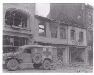 Scope and content: Original caption: American ambulance waits outside of a bombed building in Bastogne, Belgium, while a searcher looks for persons injured during the ten-day defense by 101st Airborne Div. 12/26/[44]